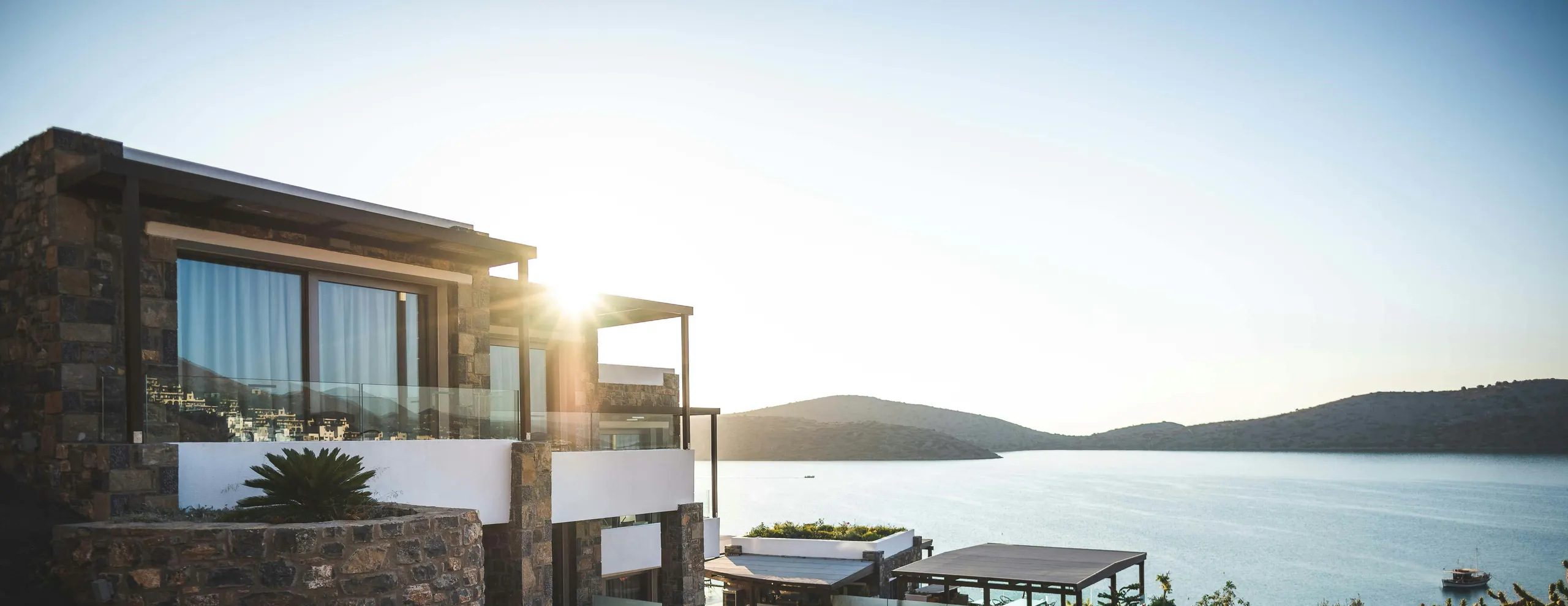 Stone-and-glass villa on a hillside above a calm bay at sunset